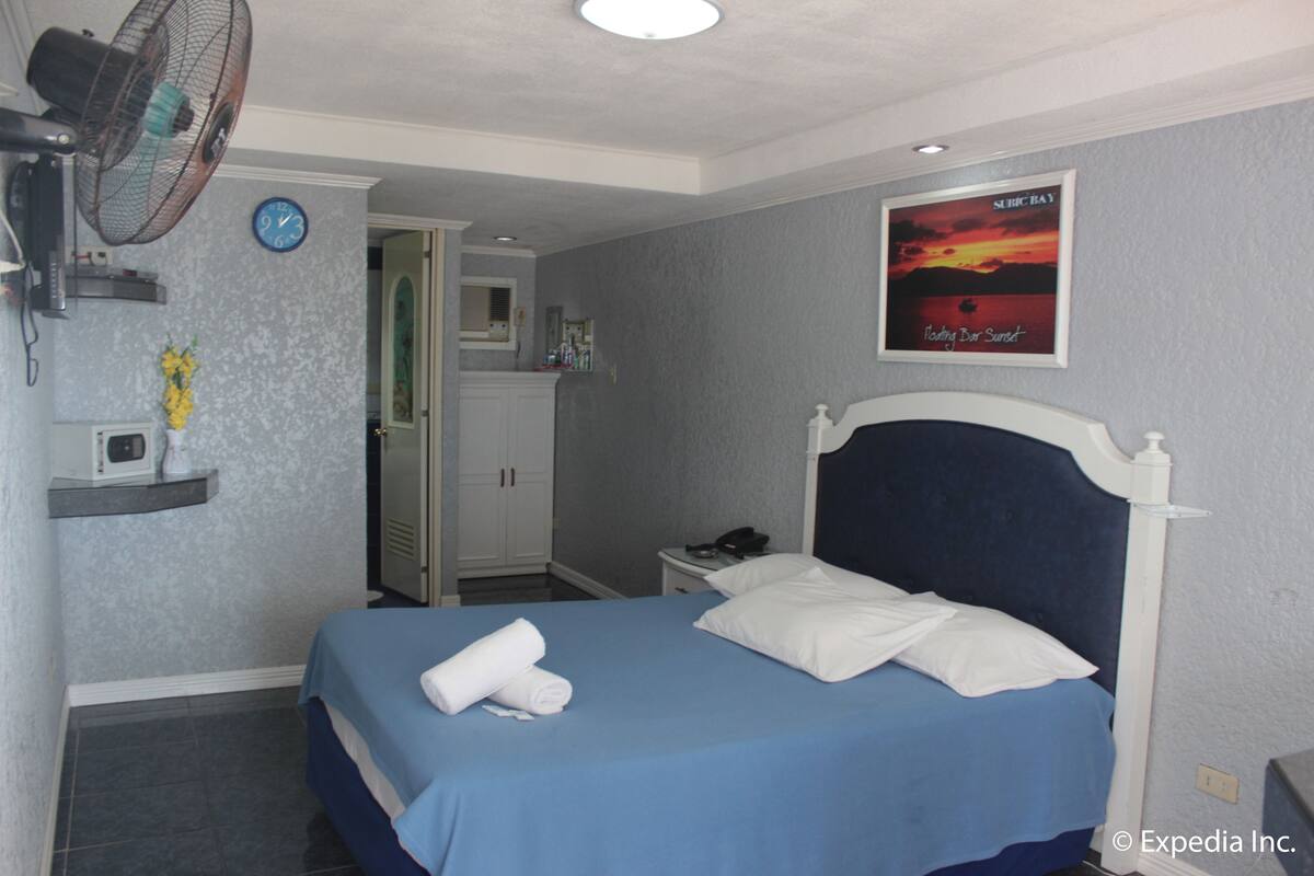 Habitación de hotel con cama, mesita de noche, ventilador, reloj, puerta del baño y un cartel en la pared.