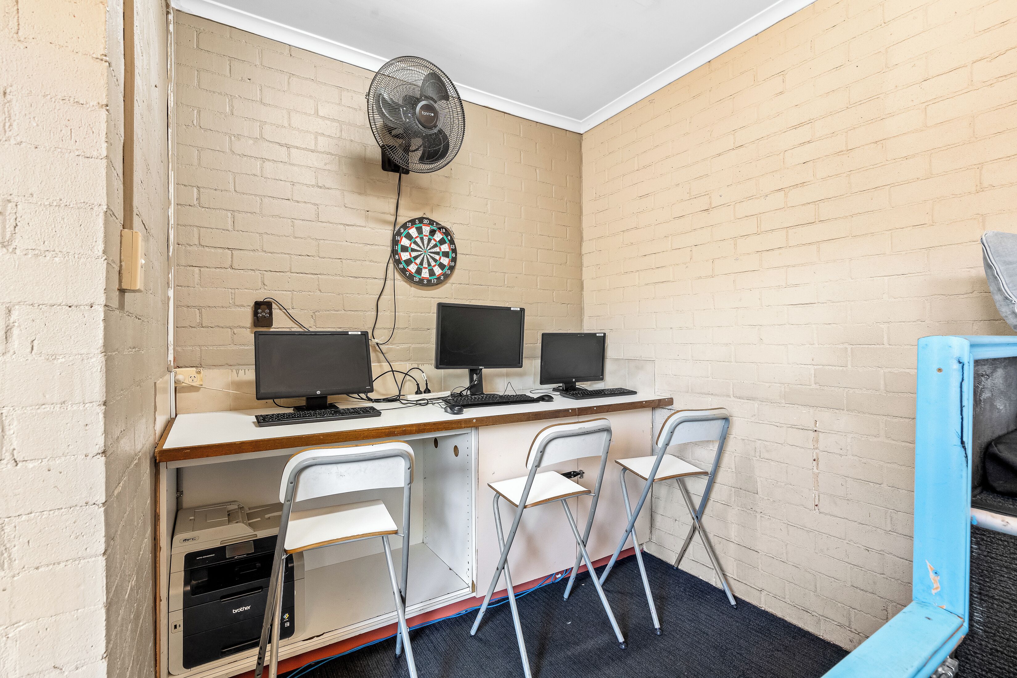 A room with a brick wall, a desk with three monitors, a dartboard, a fan, and two folding chairs.
