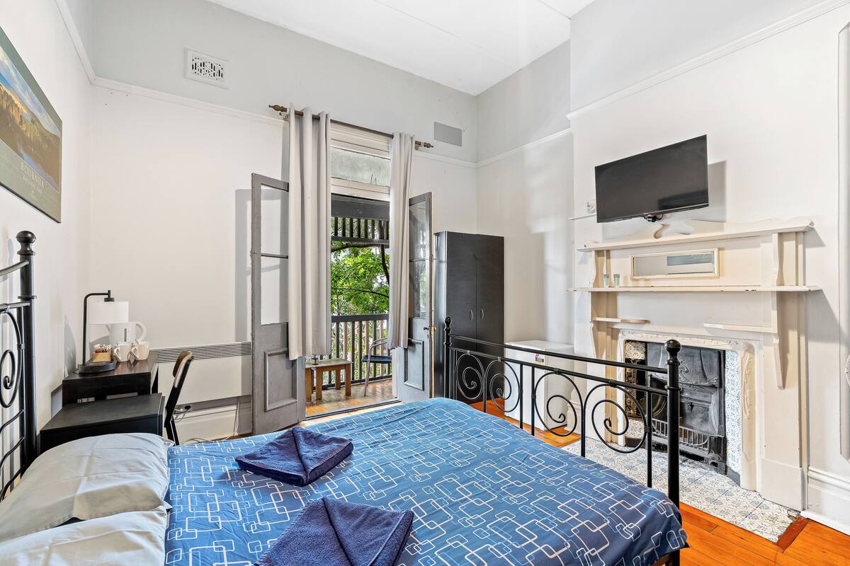 A bedroom with a bed, a desk with a lamp, a television, and a fireplace.