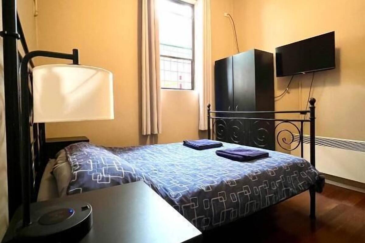 A small, compact room with a single bed, a desk with a lamp, a television mounted on the wall, and a window with curtains.