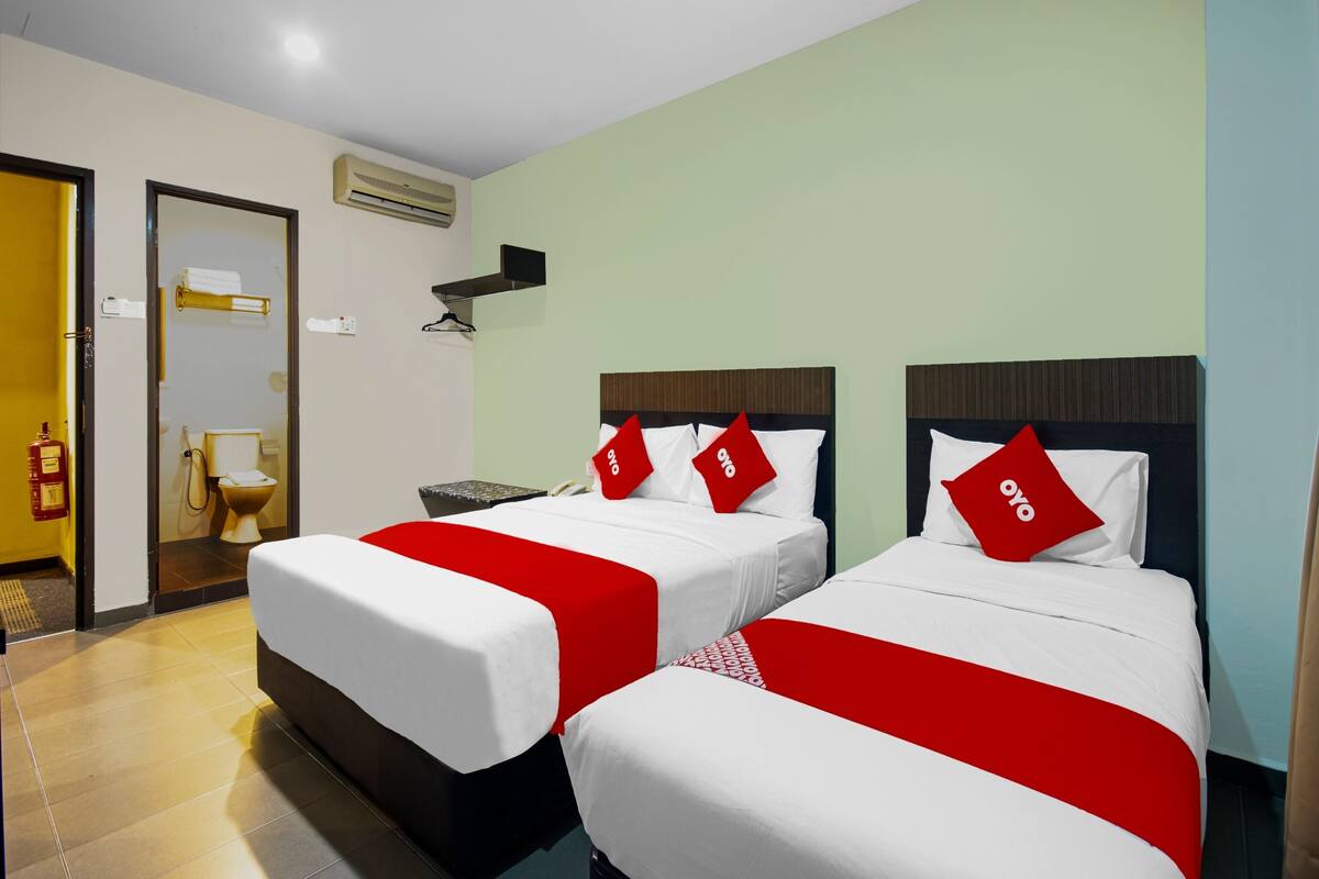 Une chambre d’hôtel avec deux lits, chacun recouvert d’une courtepointe rouge et blanche, doté d’une tête de lit en bois et orné d’un oreiller rouge portant le logo « oyo ». On aperçoit une salle de bain avec des toilettes et un porte-serviettes.