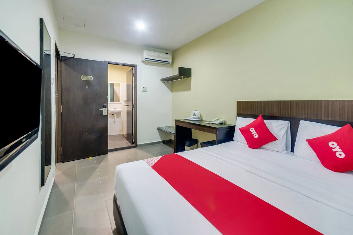 Une chambre d’hôtel avec un lit, un bureau, une chaise, un appareil de climatisation et une salle de bain visible à travers une porte ouverte.