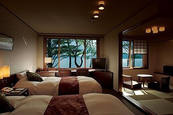 Habitación de hotel con dos camas, un escritorio con silla, televisor y ventanas amplias con vista a árboles y agua.