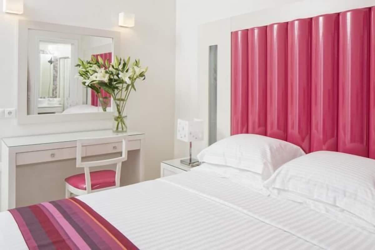 A hotel room with a bed, a desk, a chair, a mirror, and a vase with flowers.