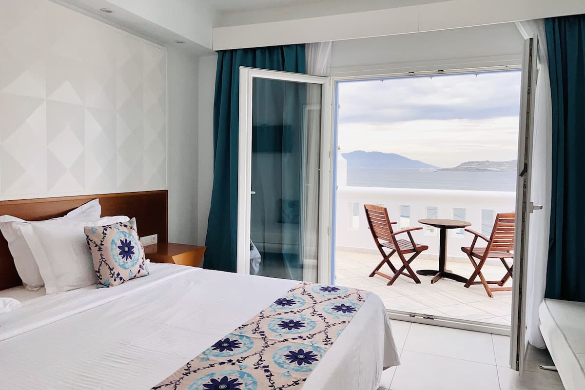 A hotel room with a bed, a nightstand, a window with a view of the sea, and a balcony with two chairs and a table.