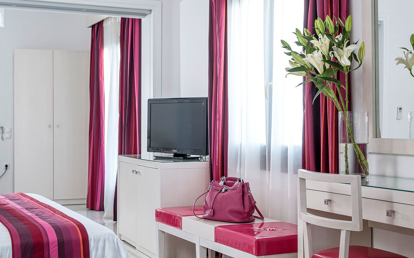 A hotel room with a bed, a television, a desk with a chair, a pink handbag, and a vase with flowers.