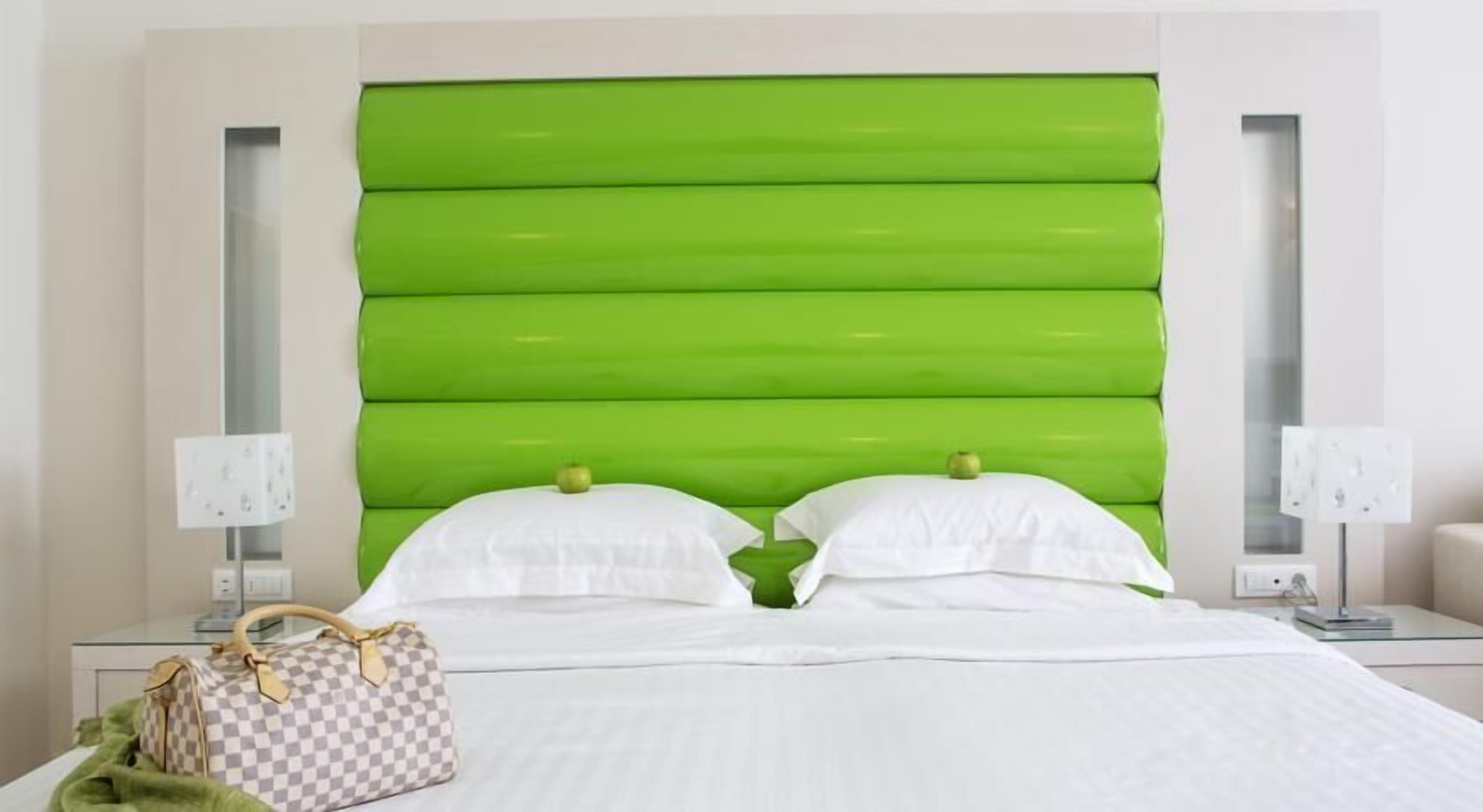 A hotel room with a green upholstered headboard, white bedding, two bedside lamps, and a checkered handbag on the bed.