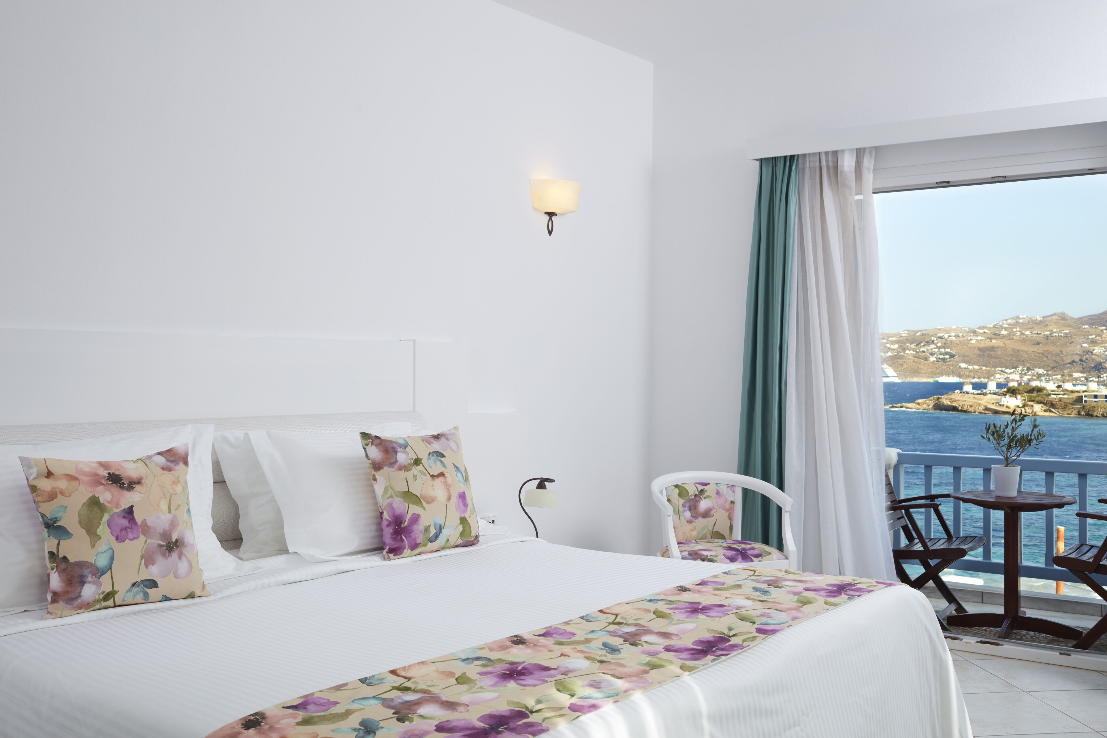 A hotel room with a bed, a nightstand, a lamp, a chair, a table, and a view of the sea.