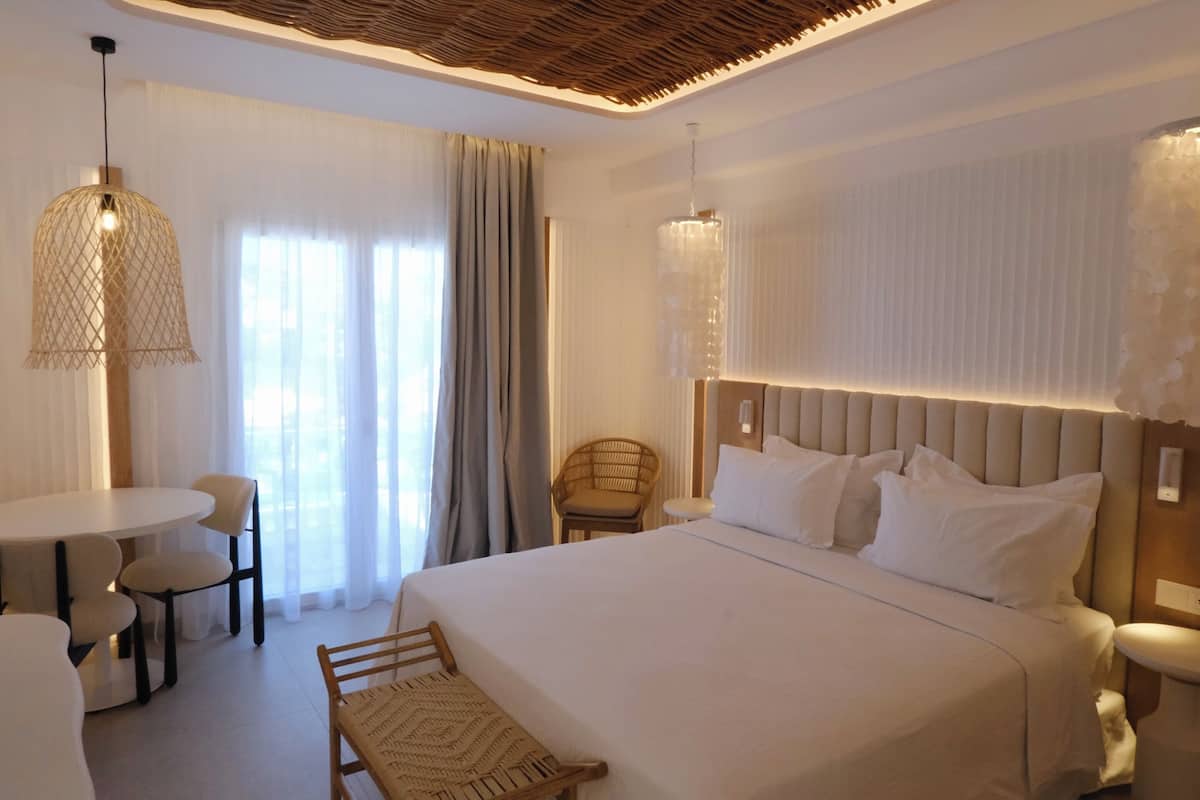 A hotel room with a bed, a dining table, chairs, a wicker chair, and a hanging light fixture.