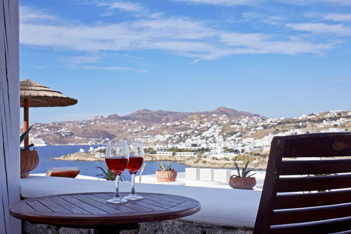 A wooden table with two wine glasses on a balcony overlooking a coastal town with white buildings and mountains in the background.