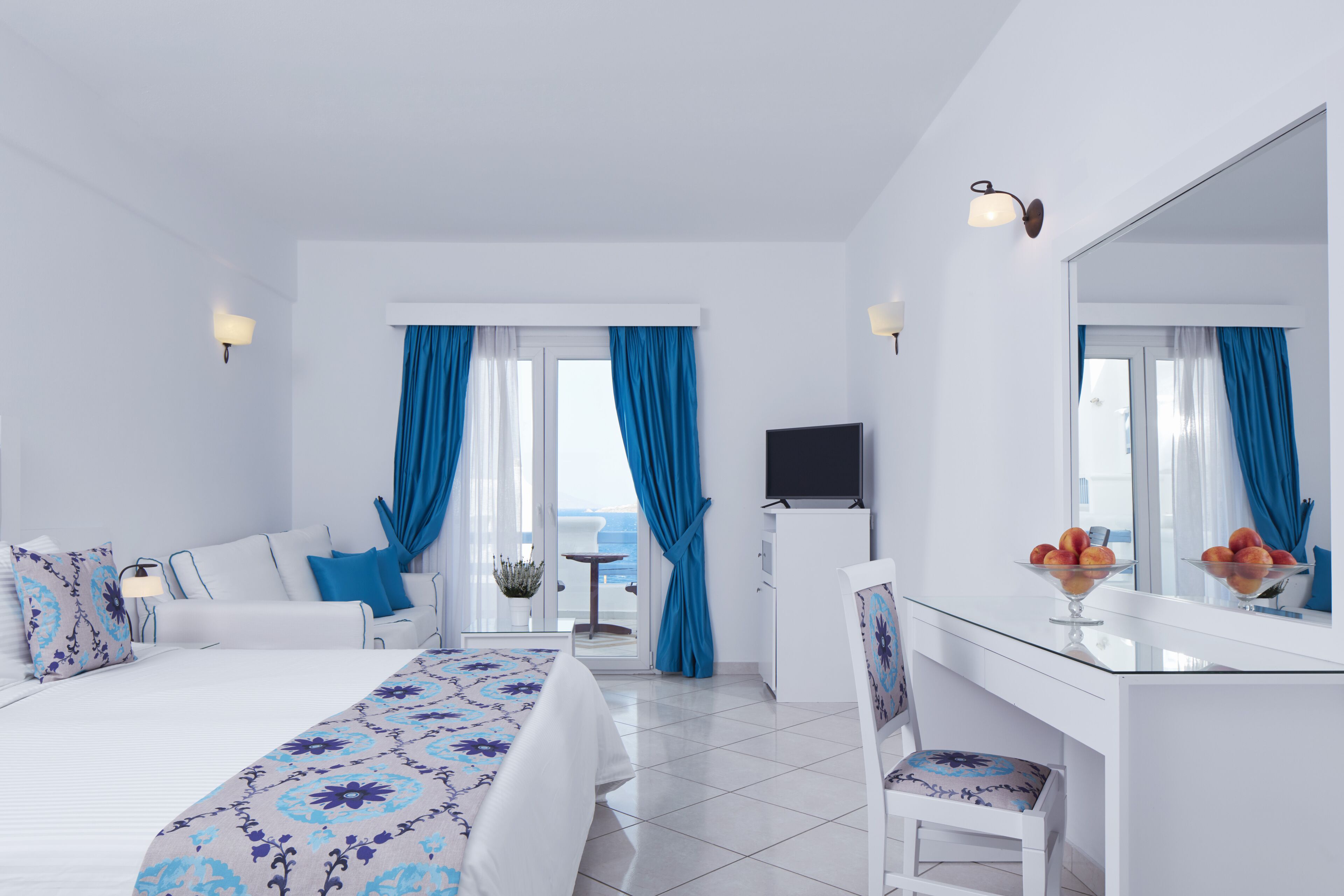 A modern hotel room with a bed, a desk, a chair, a TV, and a view of the sea.