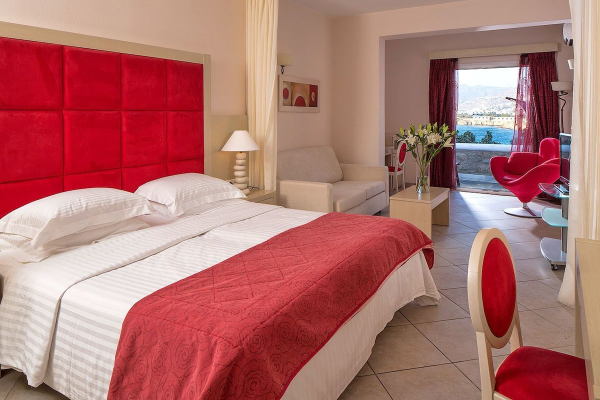 A hotel room with a large bed, a red headboard, a white sofa, a red chair, and a view of the sea.