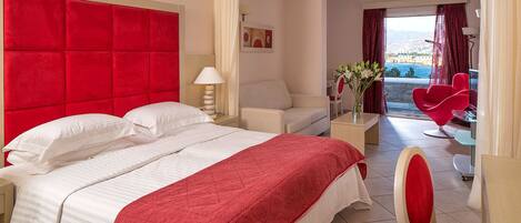 Quarto de hotel com uma cama grande, cabeceira vermelha, sofá branco, uma cadeira vermelha e vista para o mar.