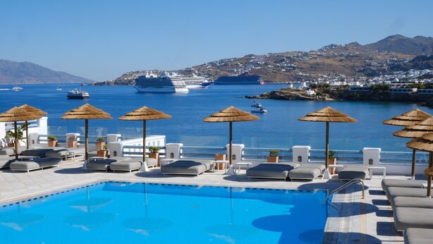 Un área de piscina con sillones de descanso y sombrillas, una piscina azul cristalina y vista al mar con cruceros al fondo.
