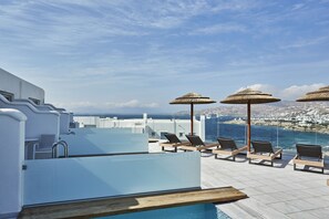 Zona de piscina en la azotea con sillones de descanso y sombrillas, con vistas a un pueblo costero.