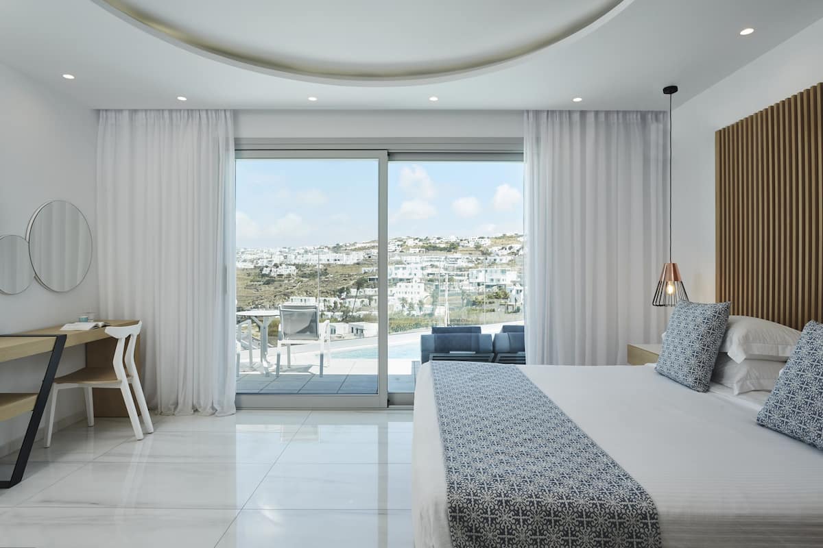 A modern hotel room with a large bed, a desk, and a view of the city through floor-to-ceiling windows.