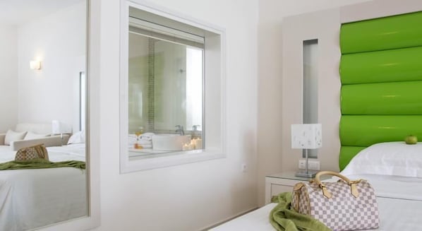 Habitación de hotel con un espejo grande, cabecera verde, cama con ropa blanca, mesita de noche con lámpara y un bolso a cuadros sobre la cama.