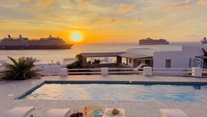 A poolside area with lounge chairs, a sun lounger, a small table with a drink, and a view of the sunset over the sea with cruise ships in the background. -  With-inn Mykonos Suites (Mykonos)