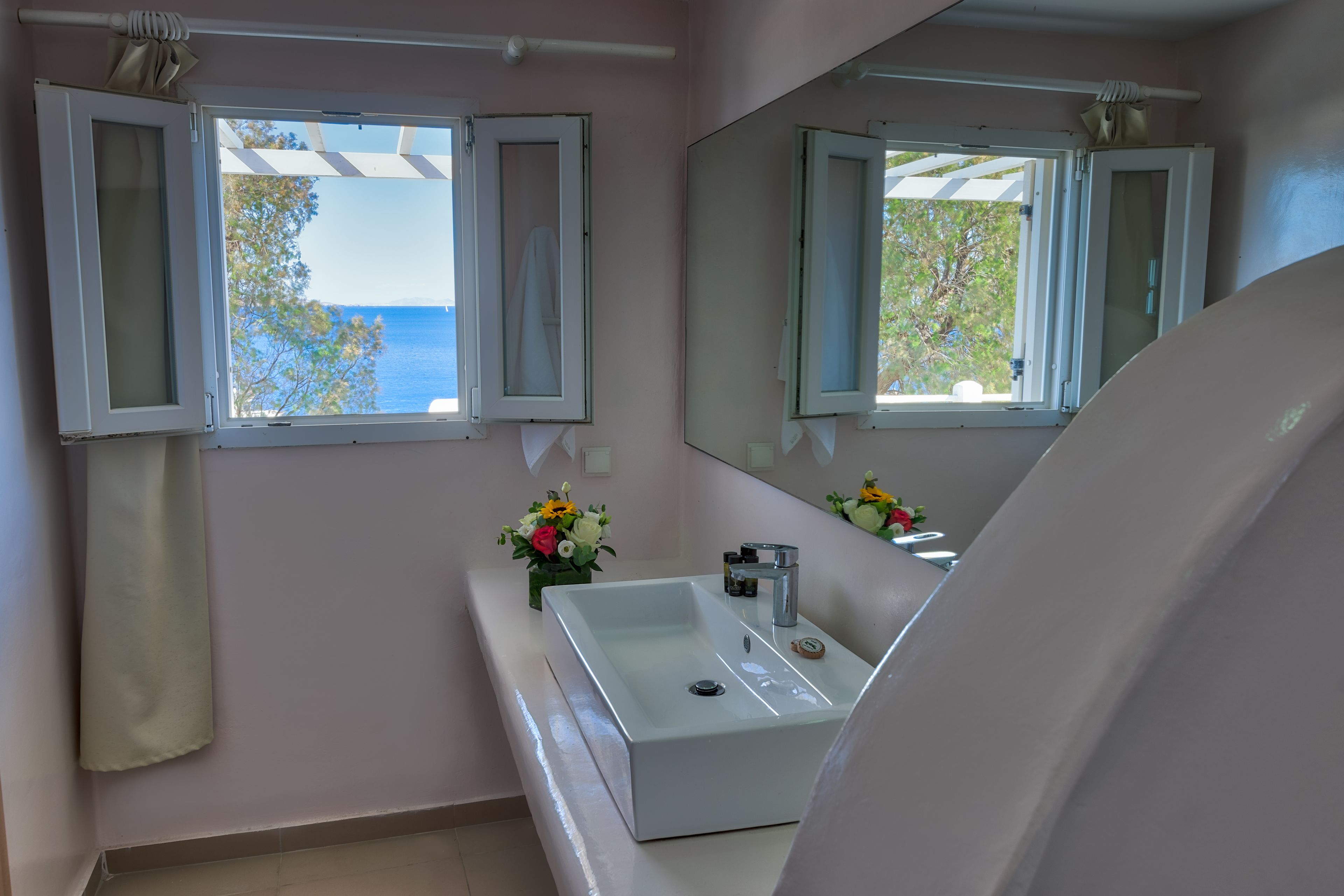 Un baño con un lavamanos blanco, un espejo y dos ventanas con persianas abiertas.