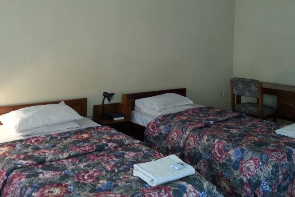 A room with two single beds, each with a floral bedspread and white pillows. There is a nightstand with a lamp and a book on it. A wooden chair with a patterned cushion is against the wall.