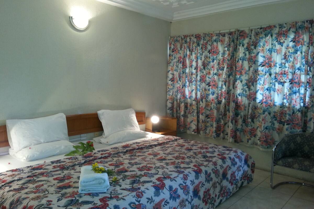 A hotel room with a bed, a nightstand, a chair, and a window with floral curtains.