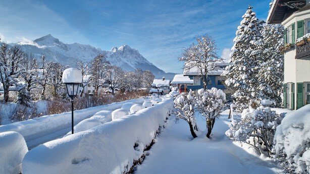 A snow-covered street with houses and mountains in the background. - Hotel Aschenbrenner (Garmisch-Partenkirchen)