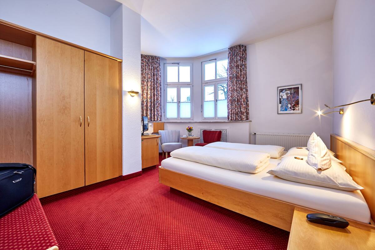 A hotel room with a bed, a wardrobe, a desk with a chair, a window with curtains, and a picture on the wall.