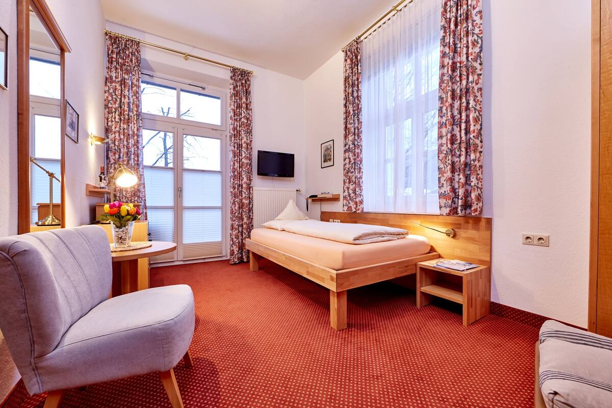 A hotel room with a bed, a chair, a small table with flowers, a TV, and curtains.