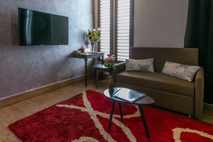A living room with a flat-screen TV, a sofa with patterned cushions, a small table with a vase of flowers, and a red and white rug.