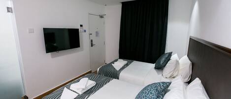 Habitación de hotel con dos camas, un televisor de pantalla plana, una puerta con perchero y un ventanal grande con cortina negra.