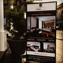 A promotional poster for the Deluxe Premier Suite at Bello Hotel, featuring a modern bedroom with a large bed, bedside tables, and a view of the cityscape.