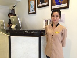 A hotel reception desk with a receptionist, a man working on a laptop, and framed pictures on the wall.