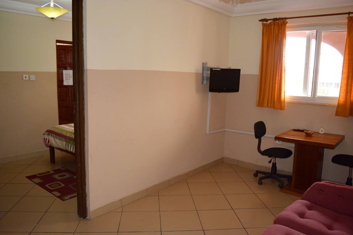 A small, well-lit room with a bed, a desk, a chair, and a television mounted on the wall.