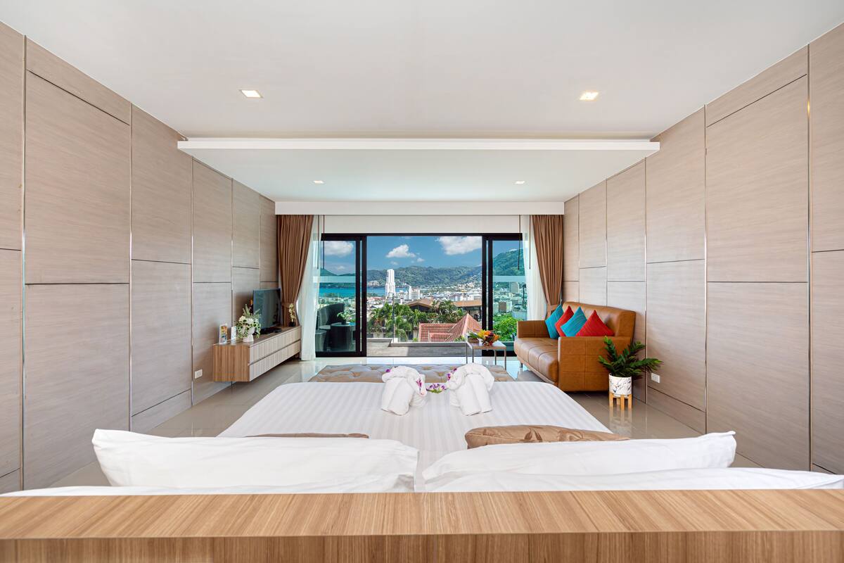 A modern hotel room with a large bed, a seating area with a sofa and armchairs, a TV, and a balcony with a city view.