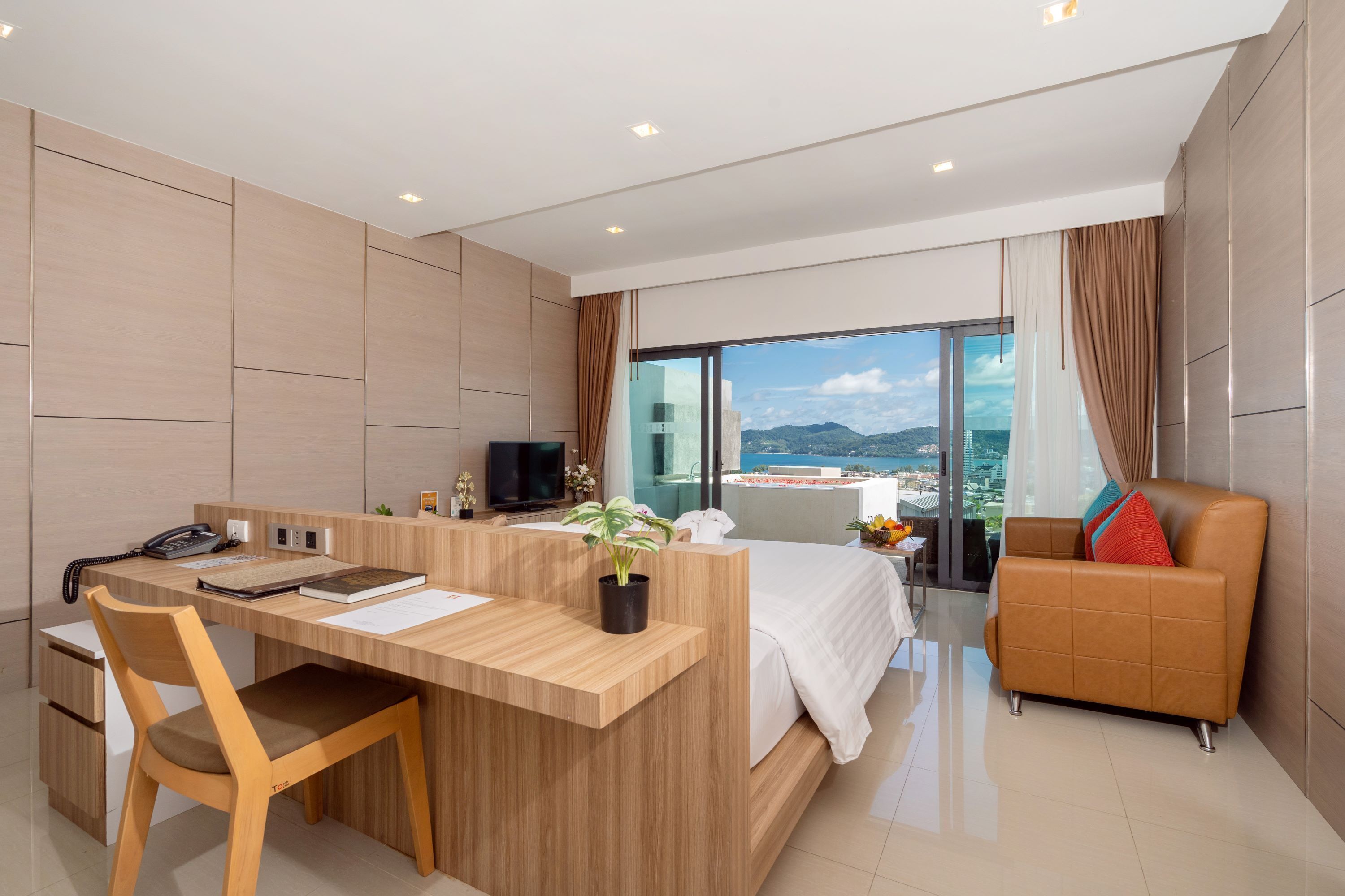 A modern hotel room with a wooden desk, a bed, a sofa, and a large window offering a city view.