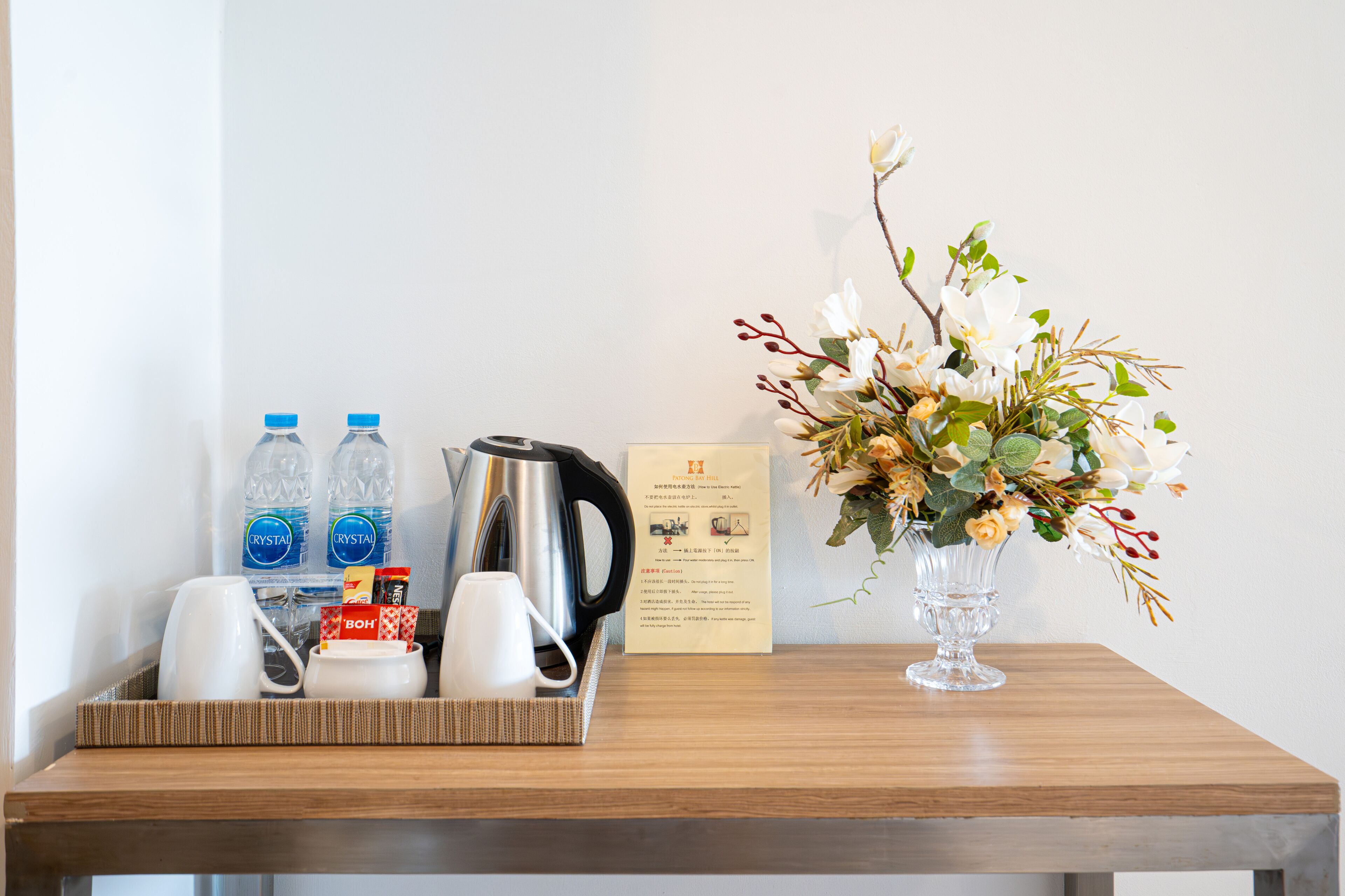 A wooden table with a tray containing a kettle, two water bottles, a coffee pot, two cups, and a small box. A vase with flowers is also on the table.