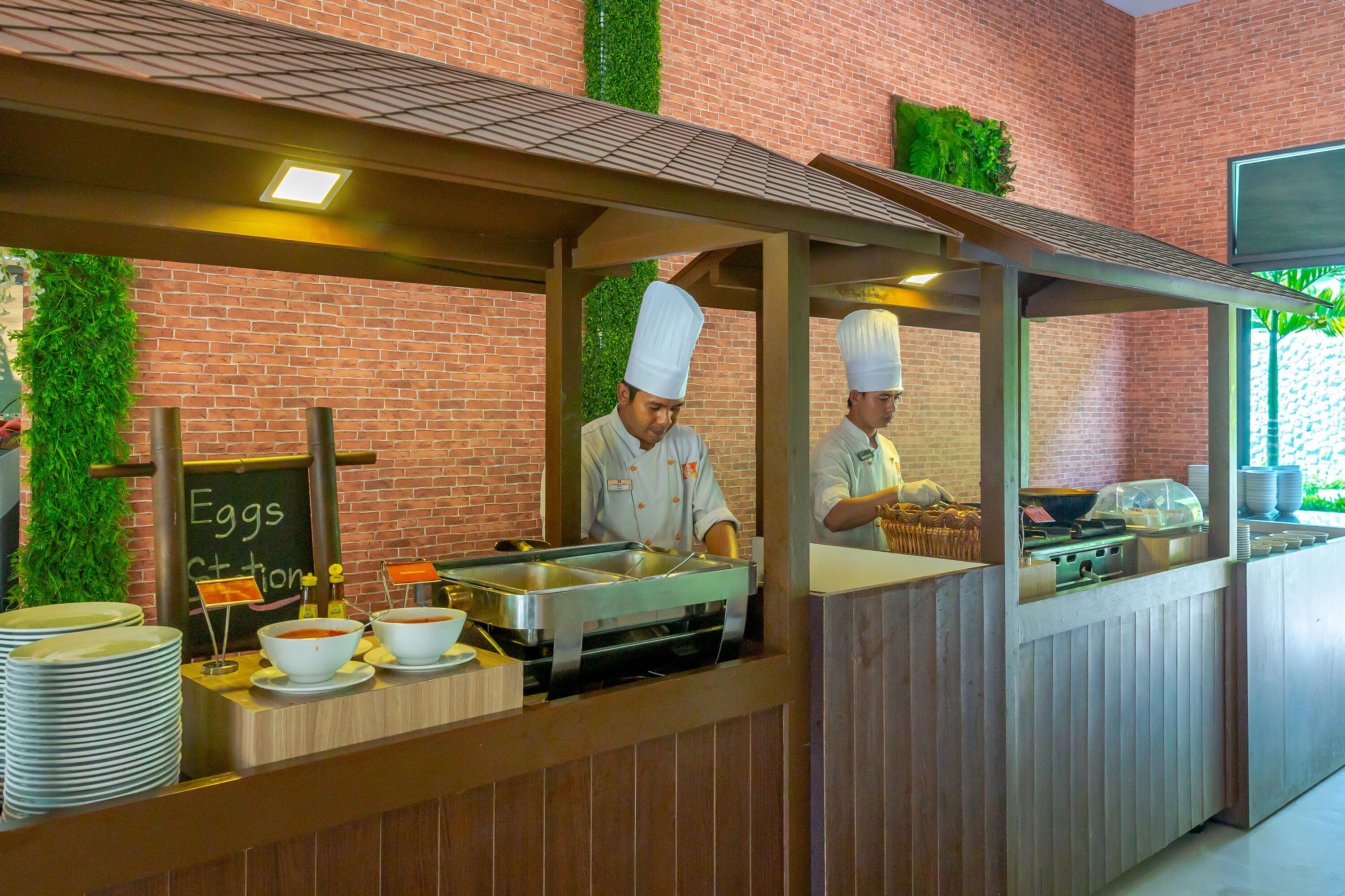 Ein Buffet mit Köchen, die Speisen zubereiten, einer Kreidetafel mit der Speisekarte und einer Backsteinwand mit Grünpflanzen.