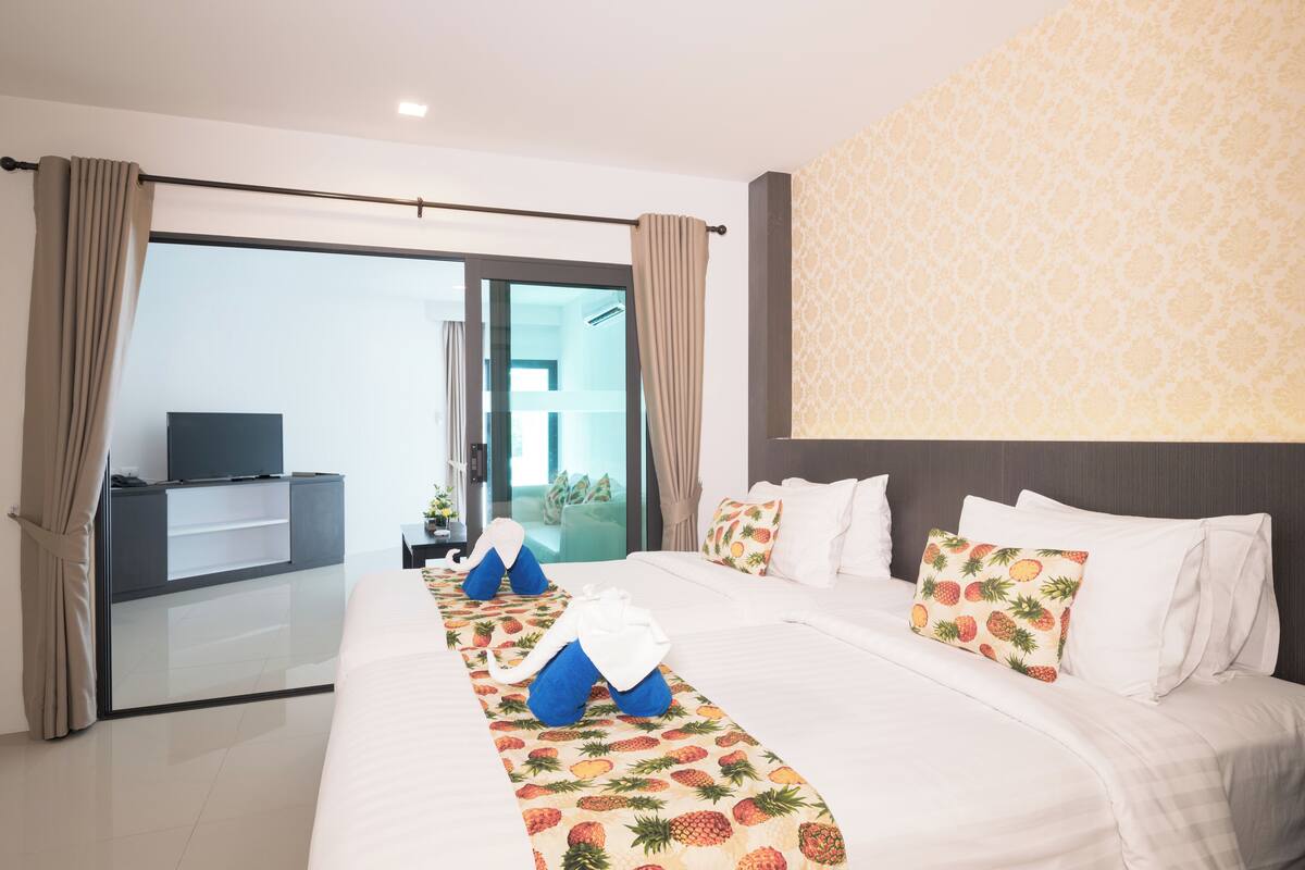 A hotel room with a large bed, a flat-screen TV on a stand, a glass door leading to a balcony, and a patterned wall.
