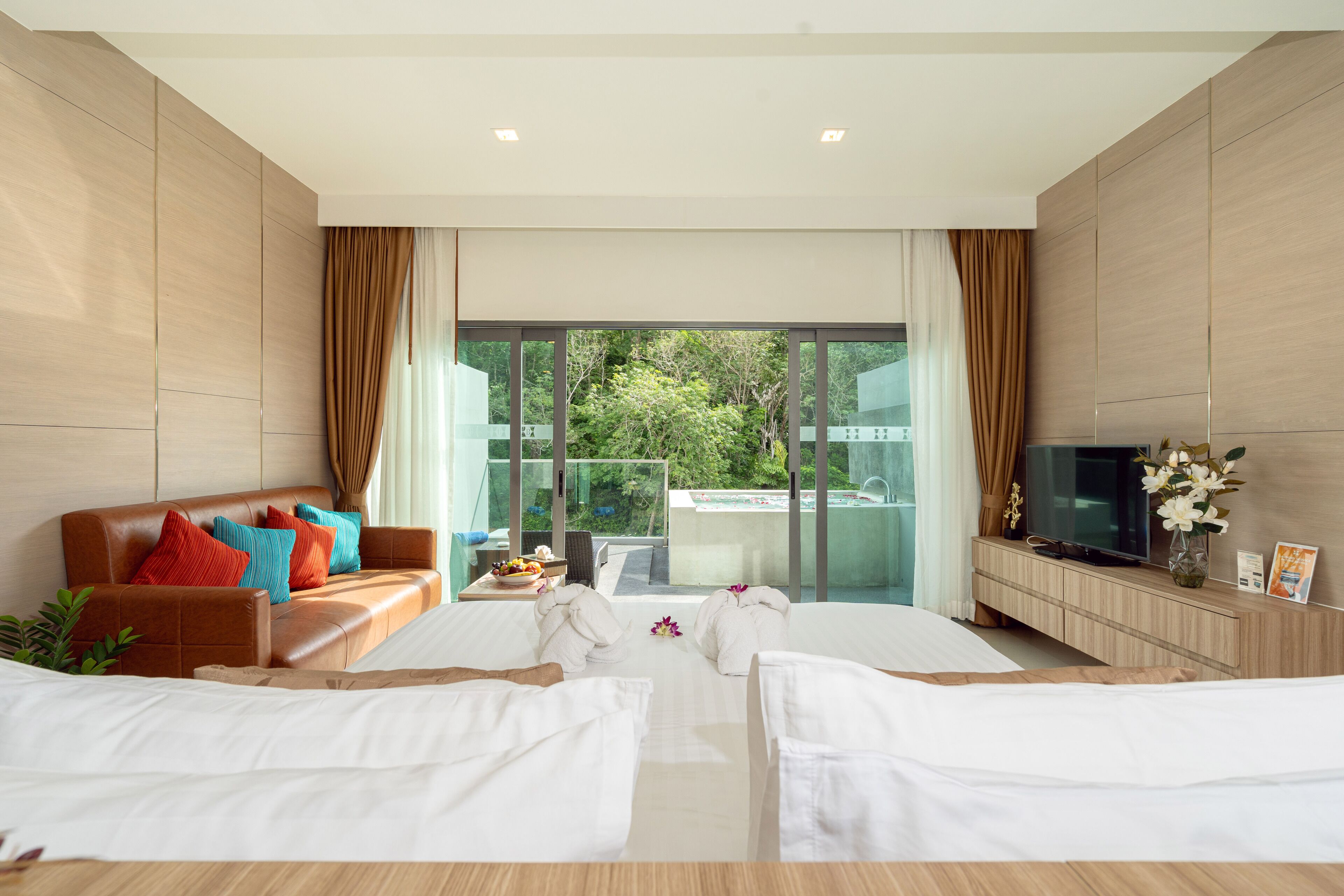 A modern hotel room with a large bed, a sofa, a TV, and a view of greenery outside.