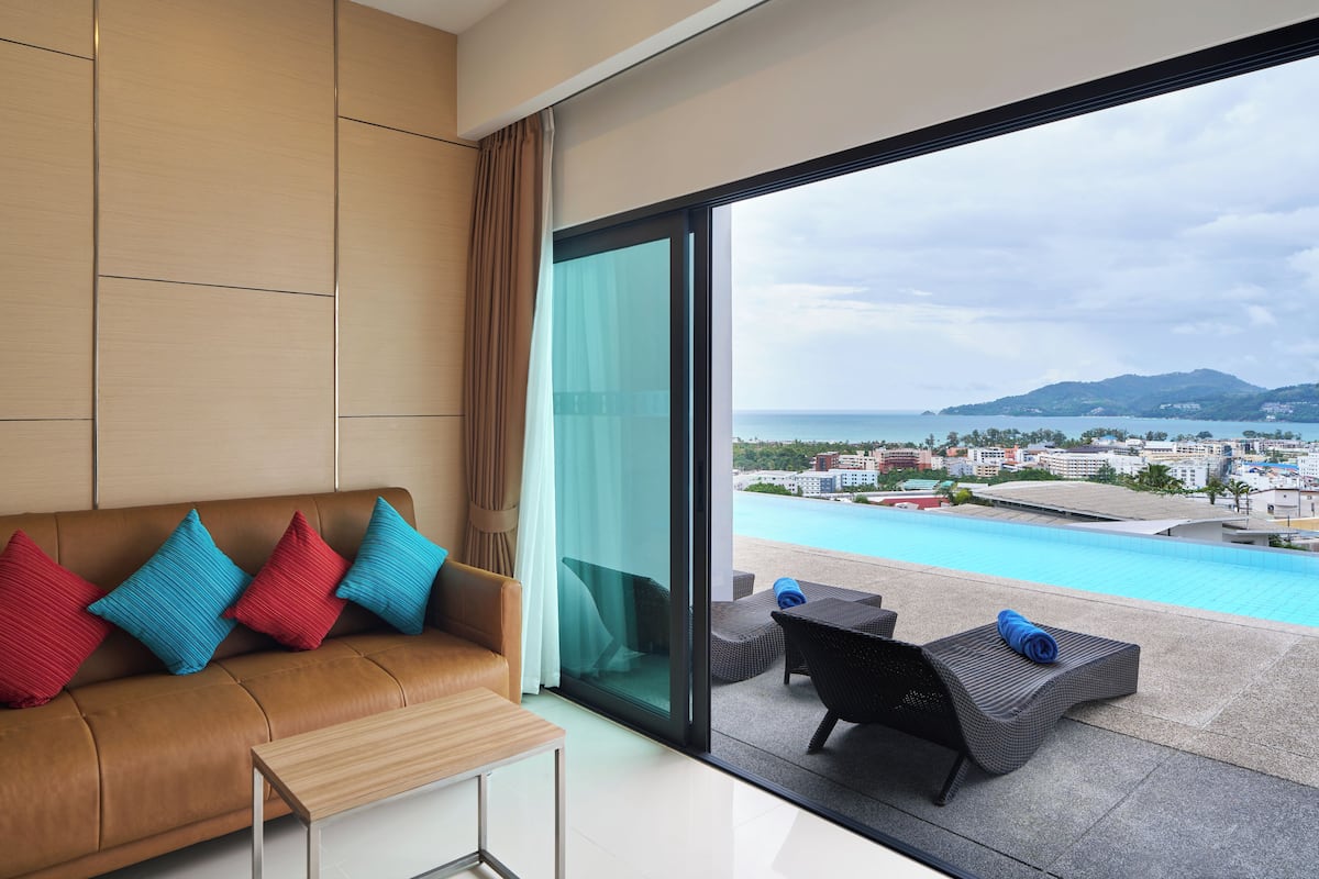 A modern room with a leather sofa, colorful cushions, a glass door leading to a balcony with a pool and city view.