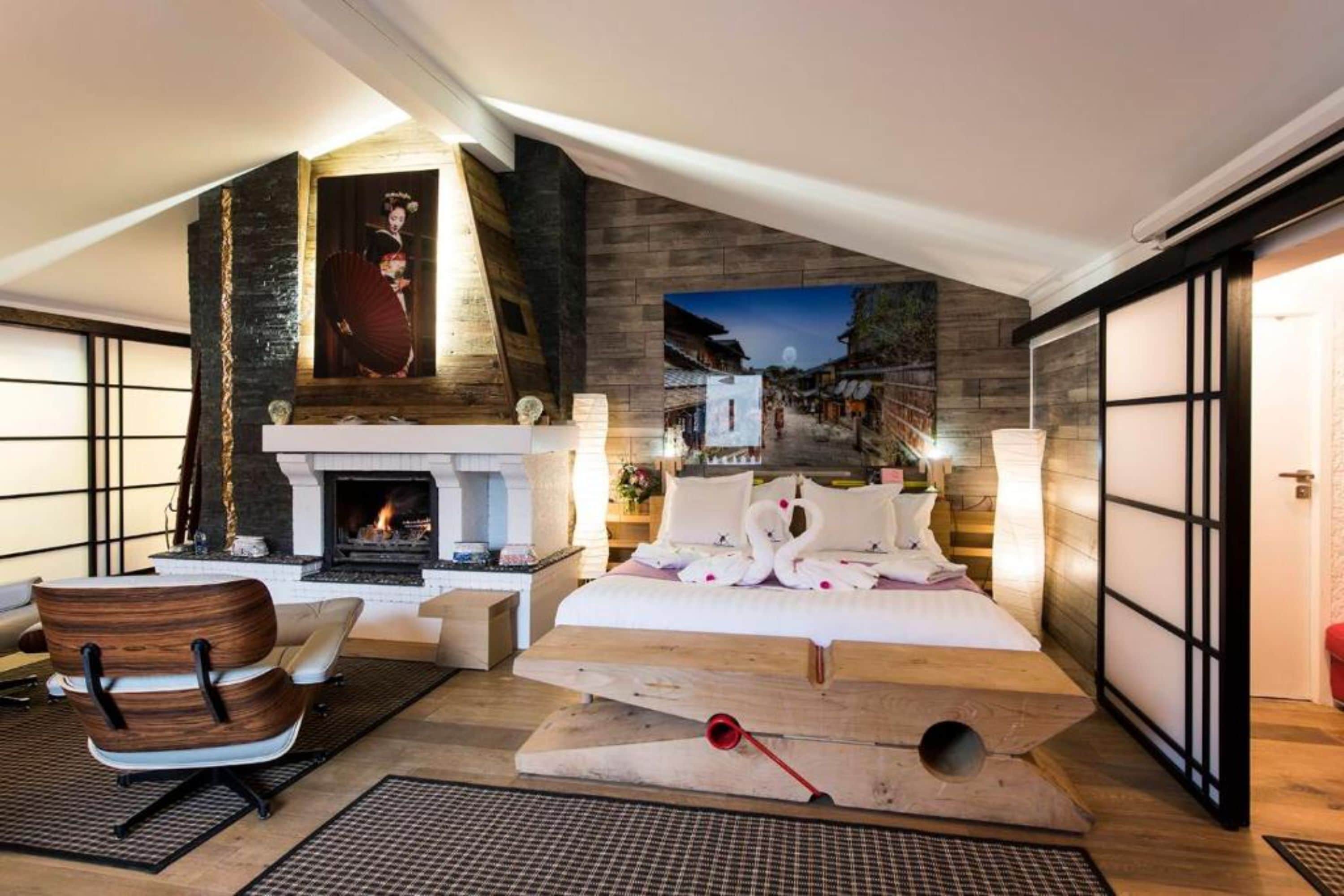 A modern hotel room with a fireplace, a bed with swan pillows, a wooden chair, and a large window with a view of a street scene.