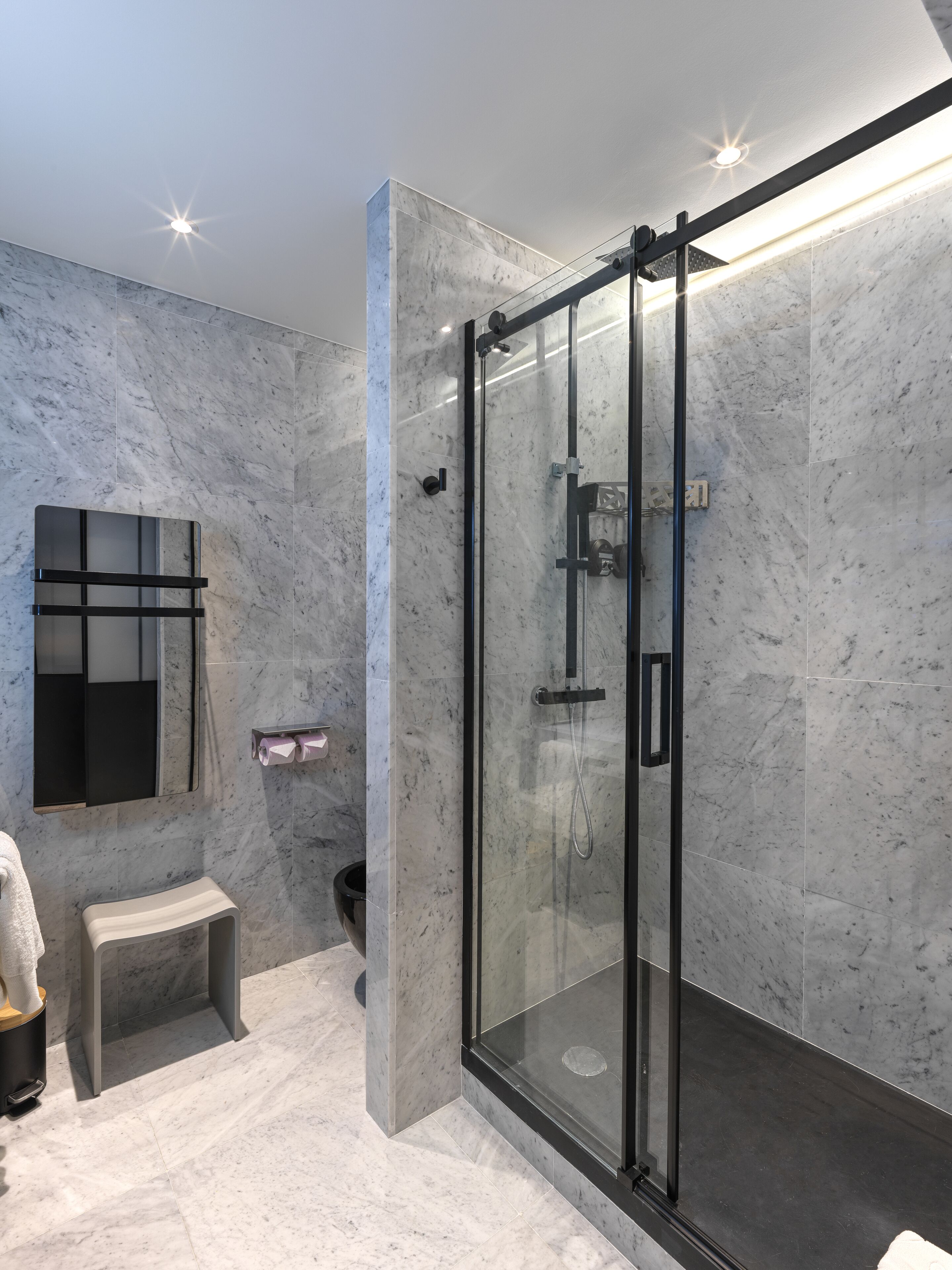 Un baño moderno con ducha envuelta en vidrio, paredes de mármol, un taburete pequeño y un perchero.