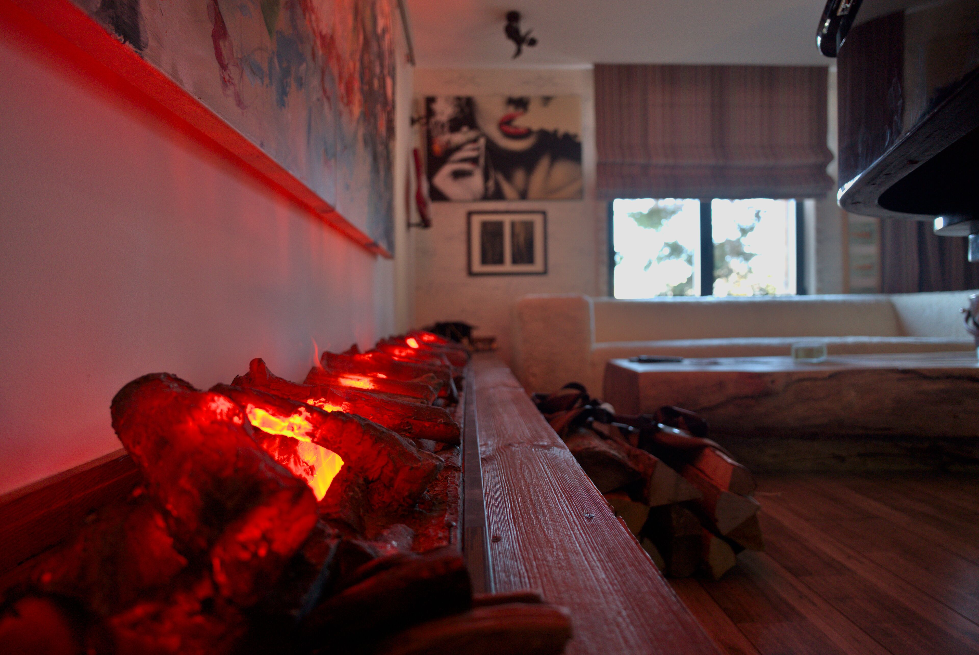 A room with a long wooden bench, a fireplace with red glowing embers, a white sofa, and a window with curtains.