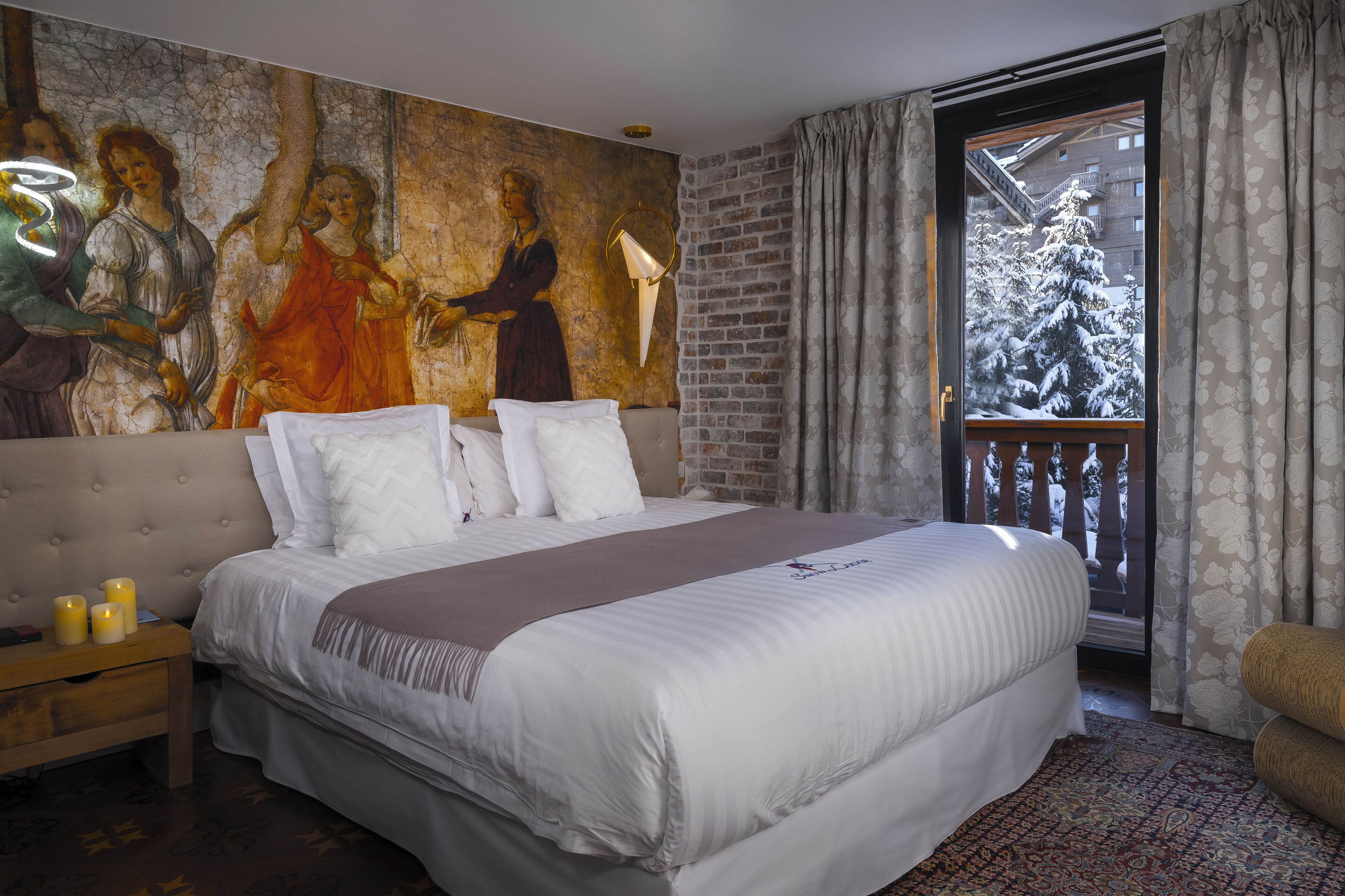 A bedroom with a large bed, a mural on the wall, a balcony with a view of a snow-covered tree, and a patterned rug.