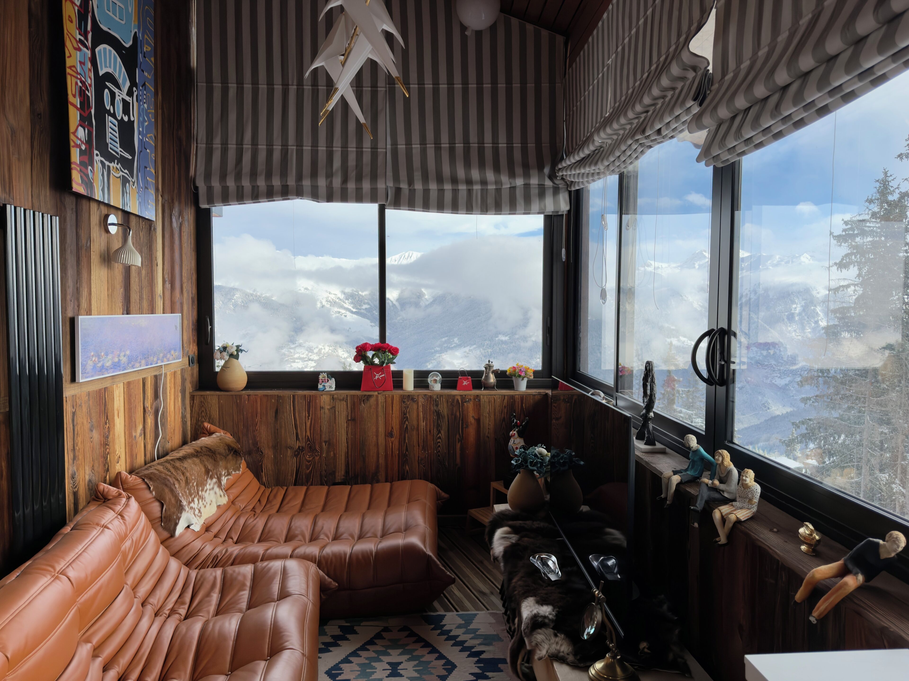 A cozy room with a leather sofa, a wooden wall, a large window with a mountain view, and a hanging light fixture.