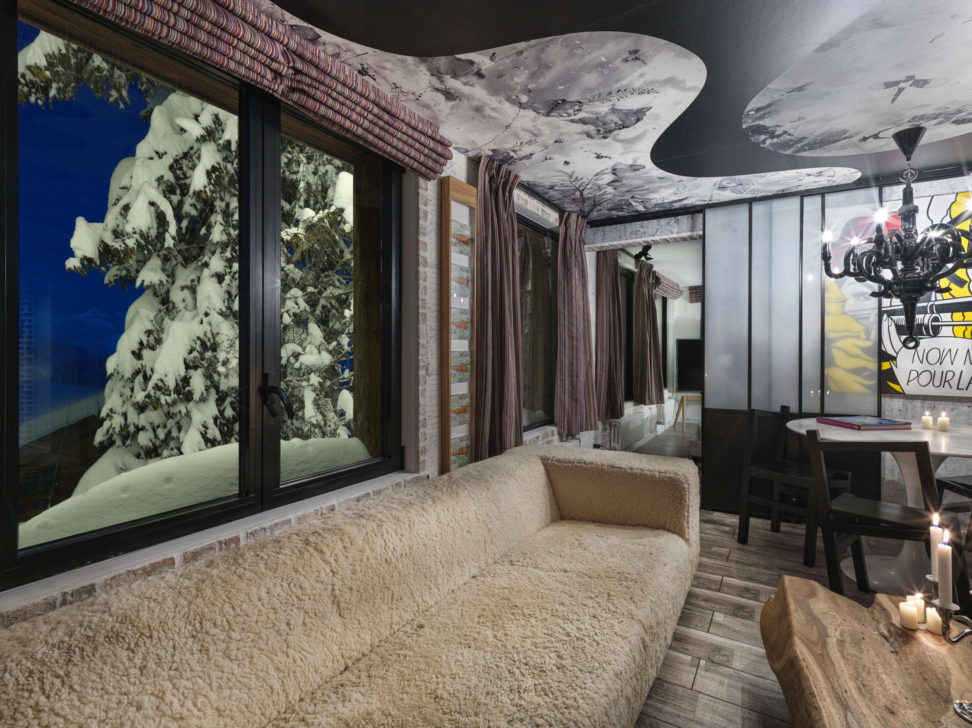 Una habitación acogedora con un ventanal grande, un sofá mullido, una mesa de comedor con velas y un mural en el techo.