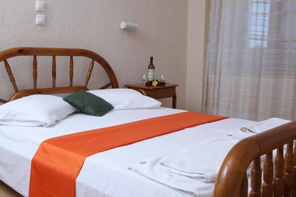 A neatly made bed with white and orange bedding, a wooden headboard, and a small bedside table with a bottle and glasses.