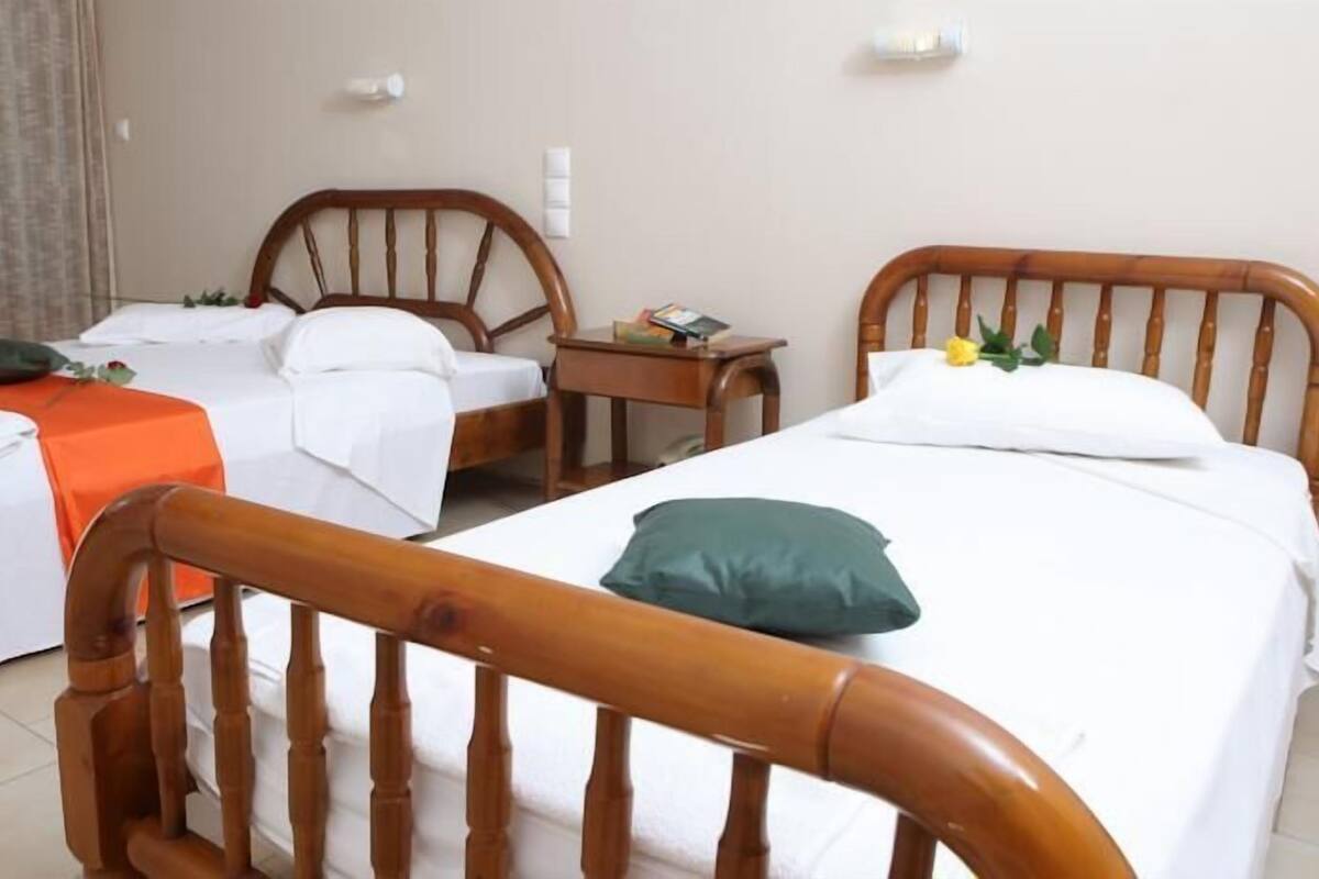 A hotel room with two single beds, each with a wooden headboard, white bedding, and a green pillow. There is a small wooden nightstand with a lamp between the beds.