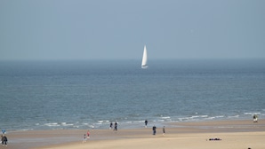 Una playa con un velero a lo lejos, gente caminando por la orilla y una extensión de arena.