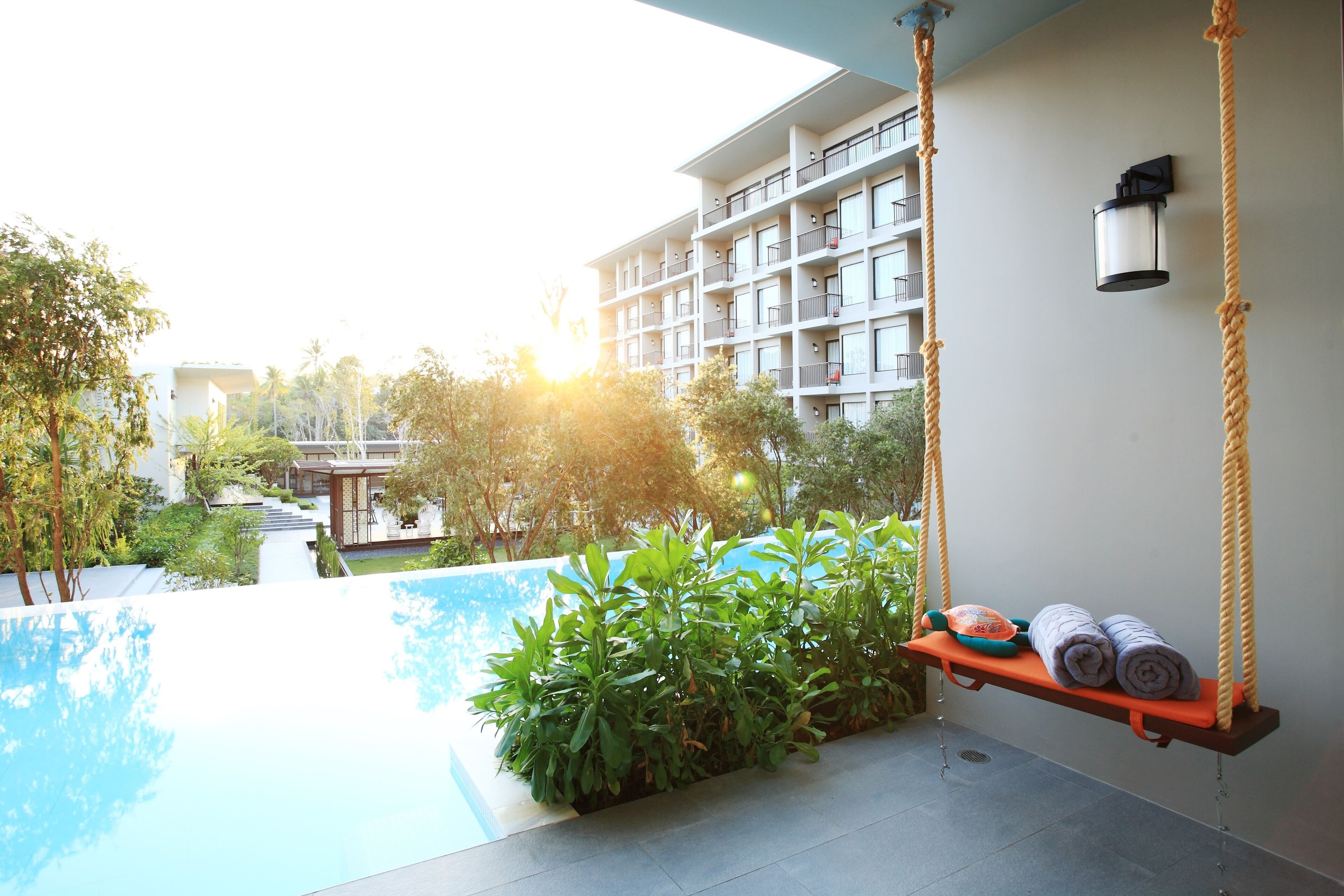 A modern hotel with a pool, a swing chair with towels, and a view of the building and greenery.